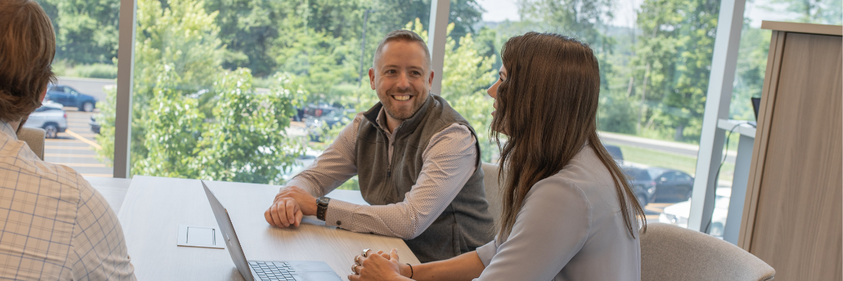 Three people sitting at a table in an office with large windows, one person using a laptop.