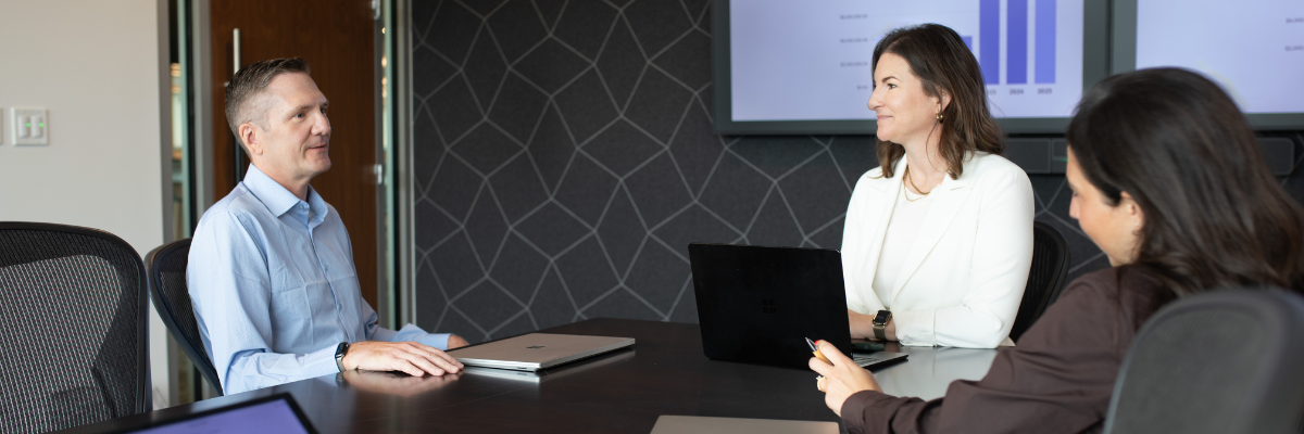 Three people are sitting around a conference table with laptops, having a meeting in an office setting with presentation screens in the background.