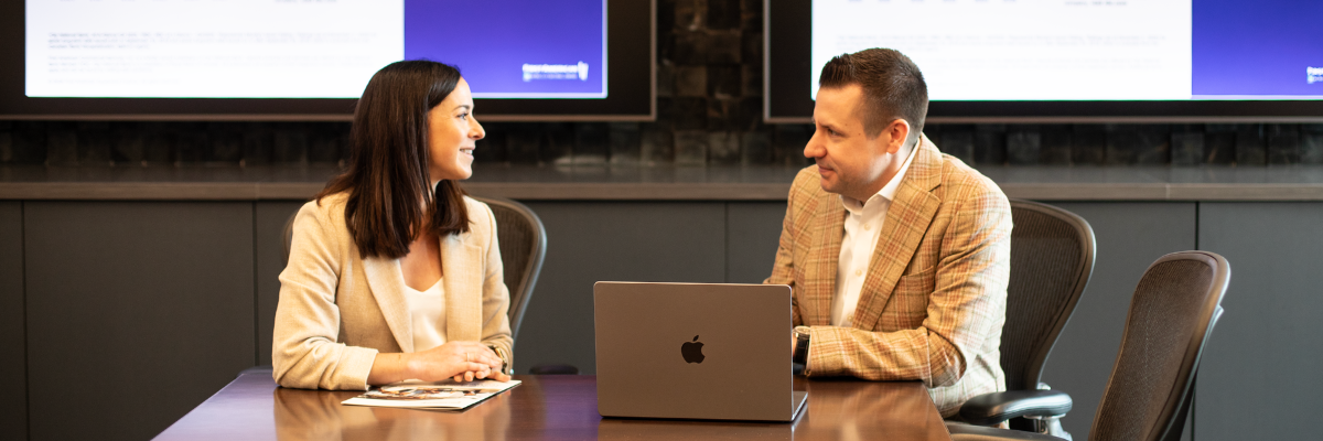 Two colleagues talk at a conference table with a laptop.
