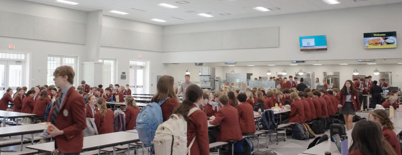 Students in matching uniforms gather and eat in a bright, modern school cafeteria with long tables and a serving area in the background.