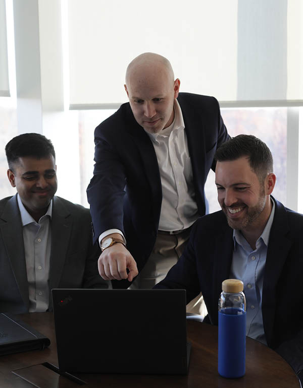 A person is pointing at a laptop screen while standing with two colleagues seated around a table in a bright office setting.