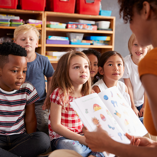 Children sit on a classroom floor listening to an adult reading a picture book.