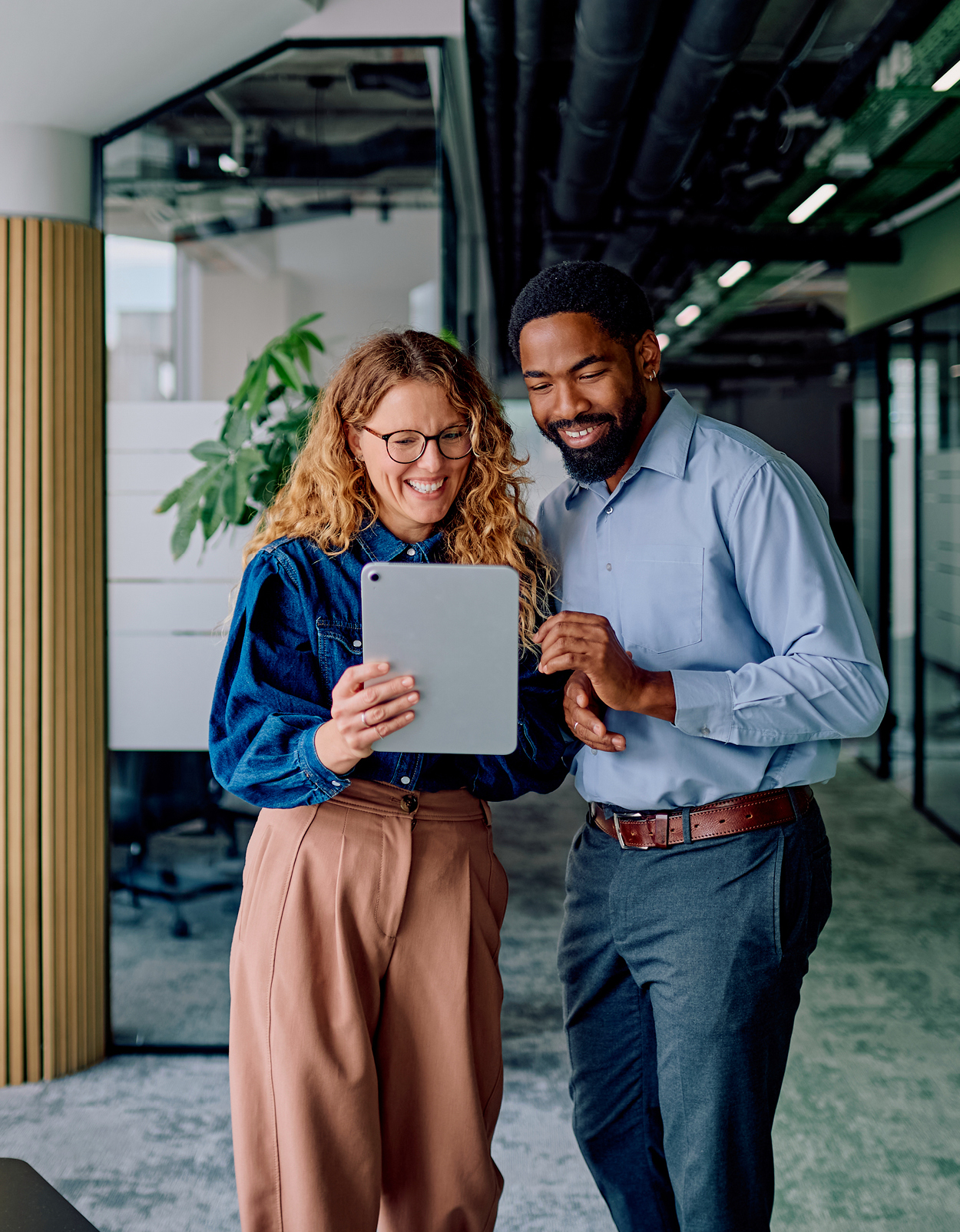 Two colleagues smiling, standing in office hallway while collaborating on tablet.