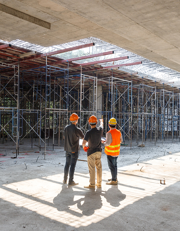 Construction professionals wearing safety helmets stand inside a building under construction, reviewing progress among scaffolding and structural supports.