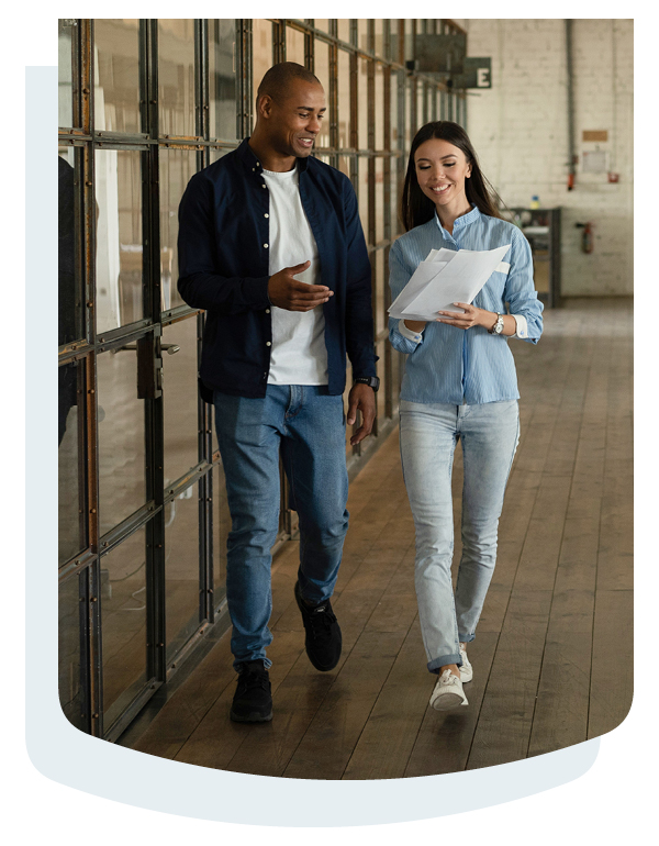 Two colleagues walking through a modern office corridor while reviewing printed documents together. Glass-walled offices line one side of the hallway, and wood flooring extends throughout the workspace.