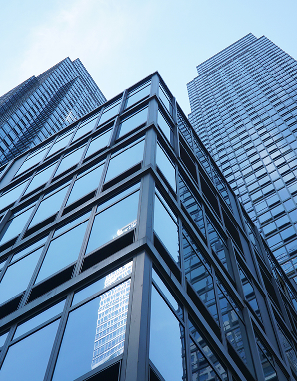 Low-angle view of several contemporary high-rise office buildings with glass facades. Reflections of neighboring structures are visible in the windows beneath a bright, cloudless sky.