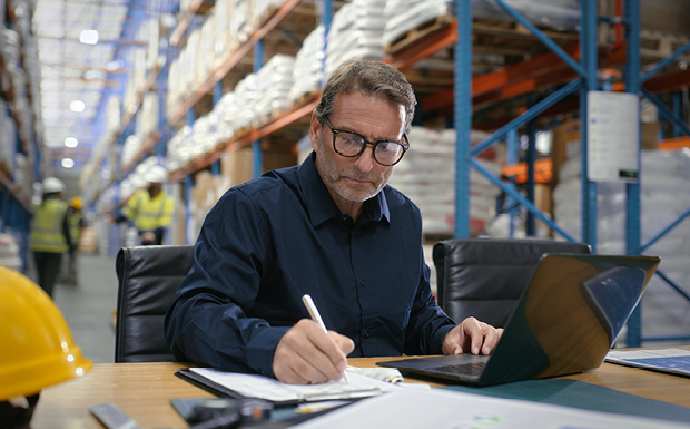 Person reviewing paperwork and working on a laptop in a warehouse.