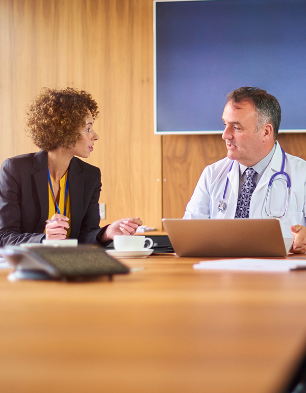 Two professionals in a conference room review information on a laptop, with one wearing a lab coat and stethoscope.