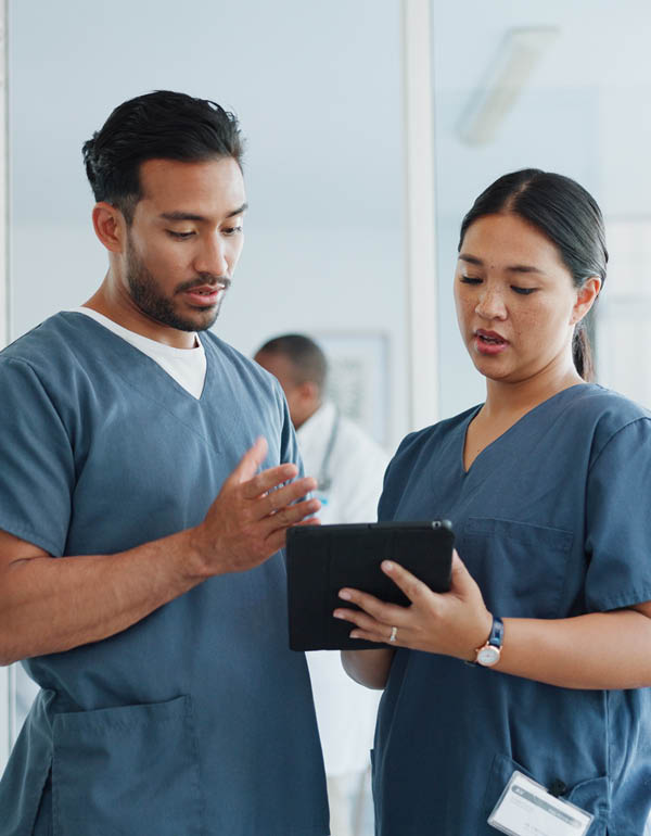 Two healthcare workers in scrubs review information together on a digital tablet in a clinical setting.