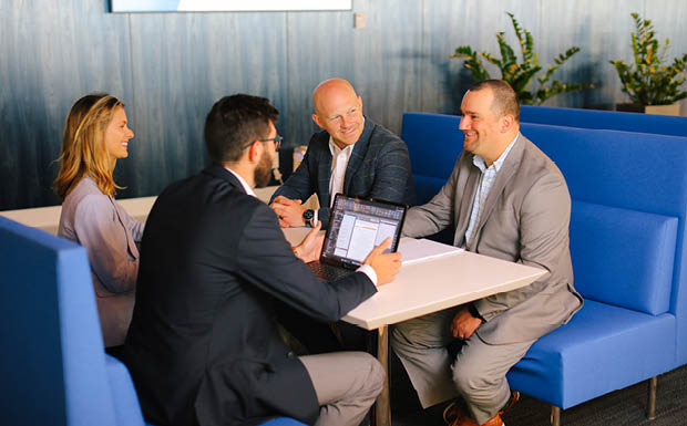 A group of business professionals sit in a booth having a discussion while reviewing information on a laptop.