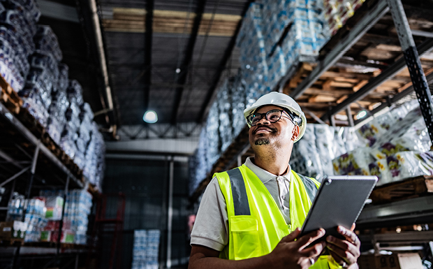 A worker wearing a high-visibility safety vest and hard hat is holding a tablet while standing in a warehouse filled with stacked goods on shelves.