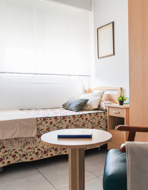 Bright bedroom with a floral single bed, small nightstand, and round table by a large window.