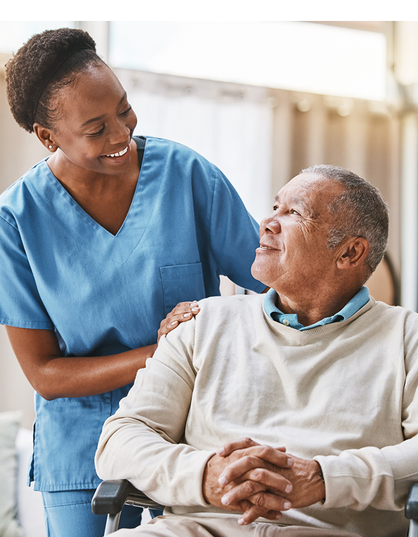 Caregiver stands beside an older adult in a wheelchair, facing each other.
