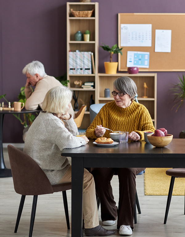 Two older adults sit at a table eating and talking in a cozy room, with another person seated in the background.