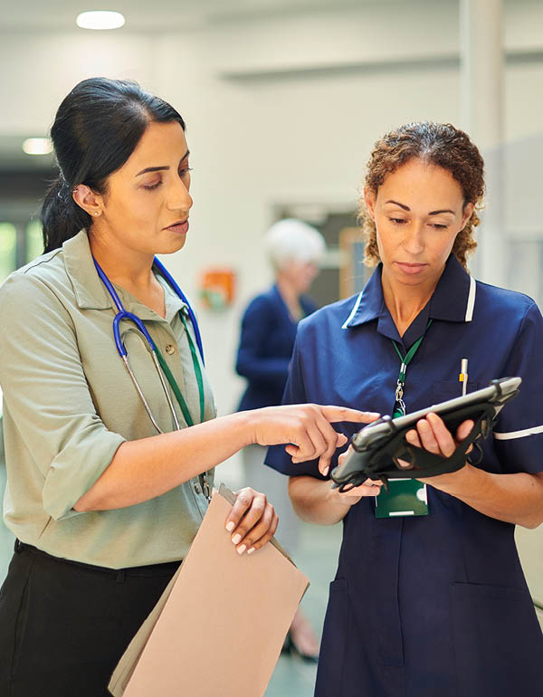 Two healthcare workers stand together in a hospital setting reviewing information on a digital tablet.