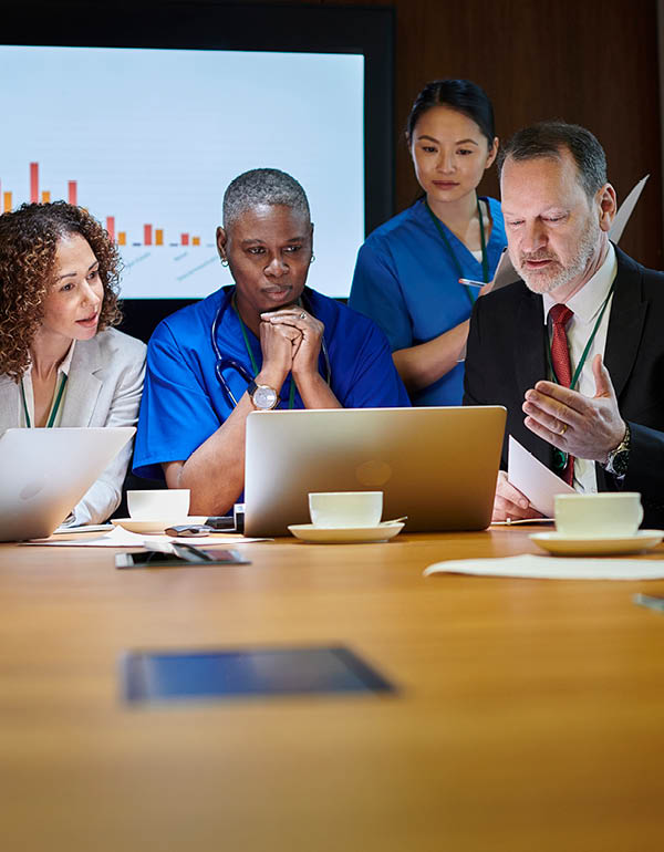 A group of healthcare and business professionals meet around a conference table, reviewing information on laptops with a presentation displayed in the background.