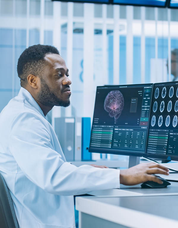 A medical professional sits at a workstation reviewing detailed brain scan images on multiple monitors.