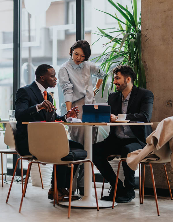 Three colleagues engage in a discussion at a modern office table, one standing and two seated.