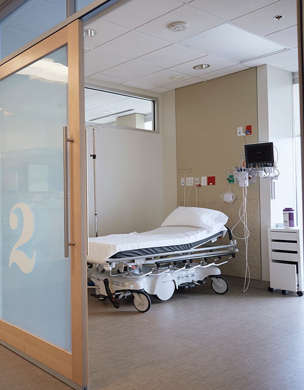 An empty hospital patient room with a bed, monitoring equipment, and a glass door labeled with the room number.