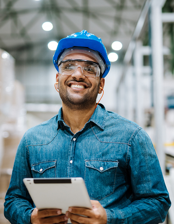A person wearing a blue hard hat and denim shirt holds a tablet while standing in an industrial warehouse setting.