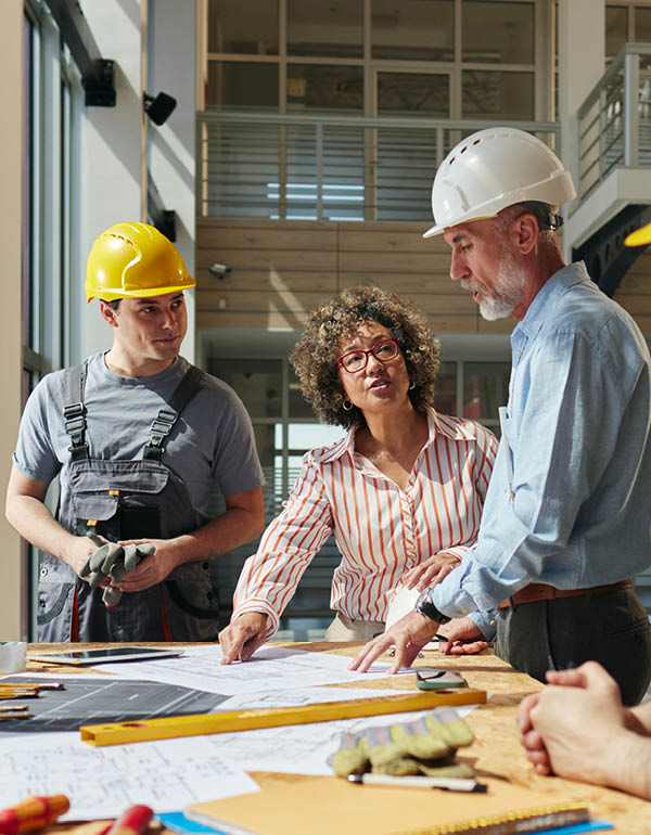 Three individuals, including two in hard hats, collaborate over architectural plans in a modern workspace, representing construction or engineering planning.