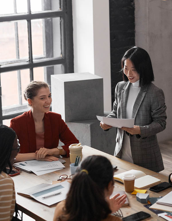 Four people in a modern office meeting; one stands with papers, others sit with documents and coffee.