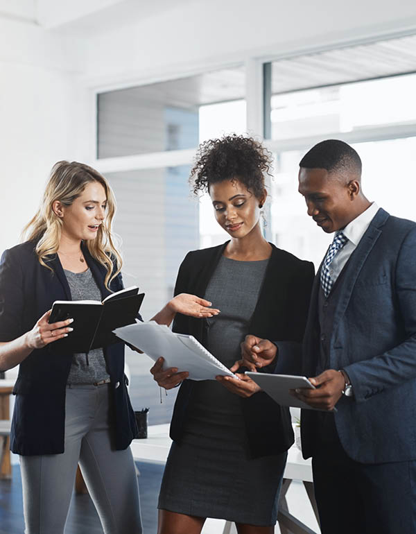 Three professionals in business attire discuss documents and a tablet in a collaborative office setting.