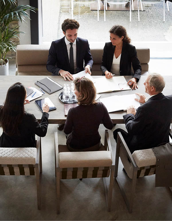 Five people sit around a large table in a business meeting, reviewing documents and taking notes.