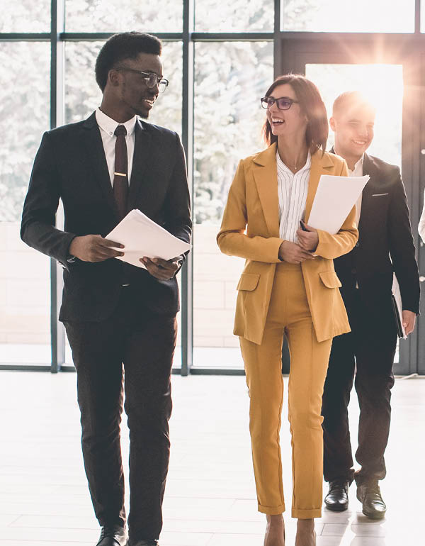 Two professionals walk through a bright office hallway, conversing and carrying documents.