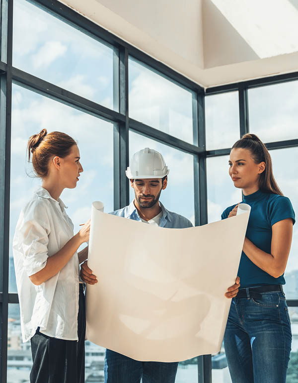 Three people review blueprints in a bright room; one wears a hard hat.