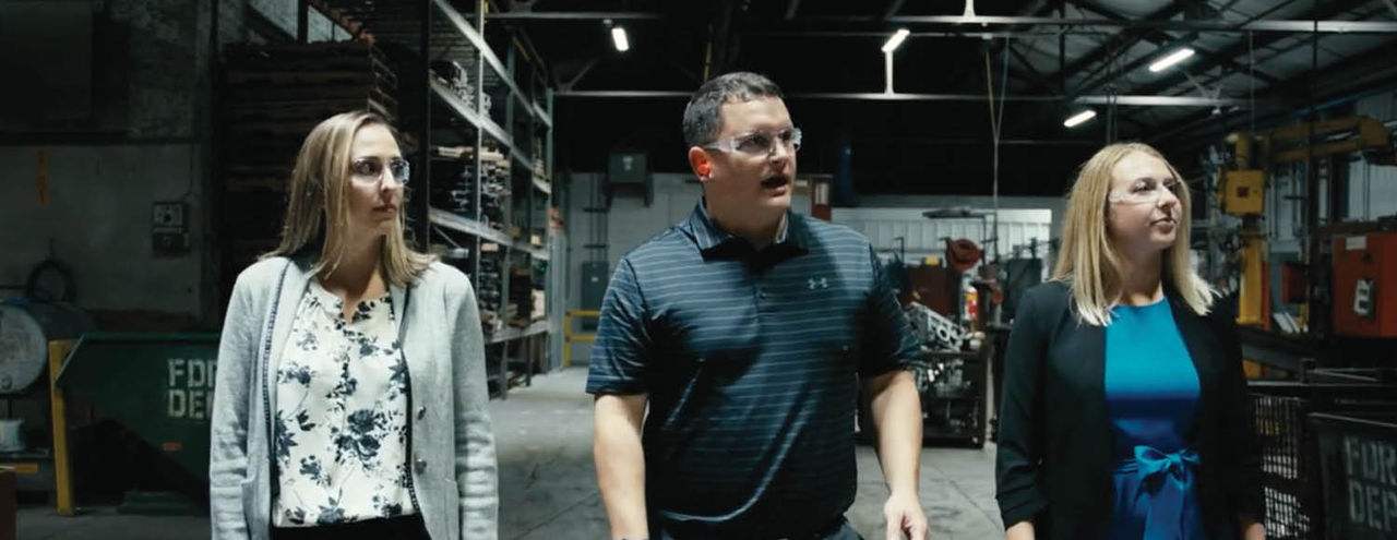 Three people walk together through an industrial warehouse filled with metal shelving, machinery, and overhead lighting.