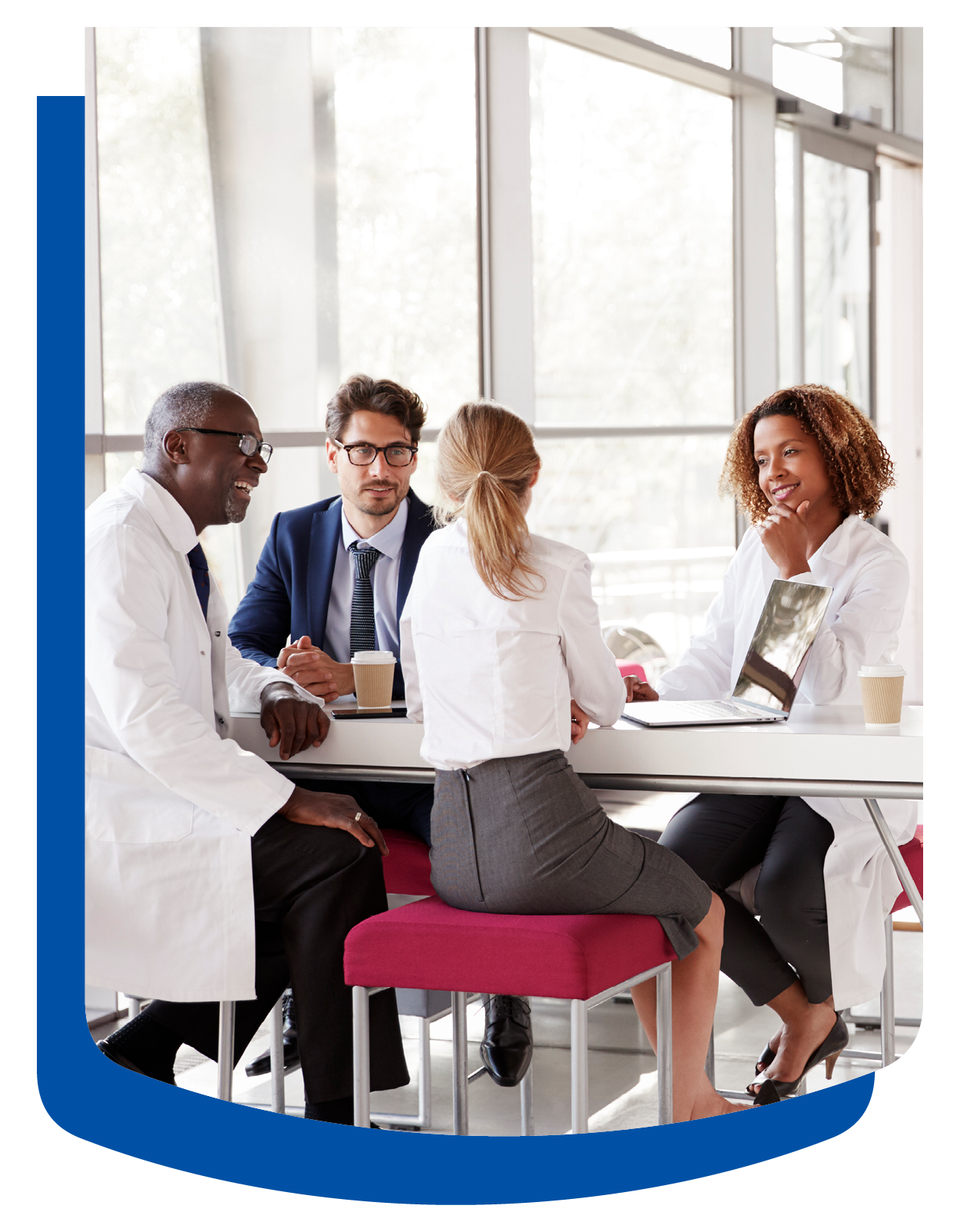 A group of professionals in a bright office setting sit around a high table with a laptop and coffee, engaged in a collaborative discussion.