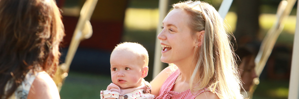 A woman holding a baby outdoors while talking to another person in a sunlit setting.