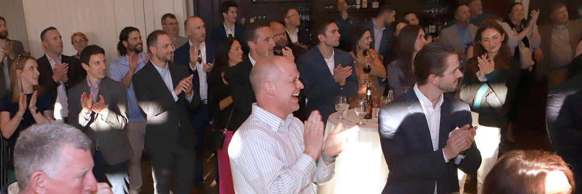 A group of business colleagues standing closely together and clapping in a softly lit indoor venue.