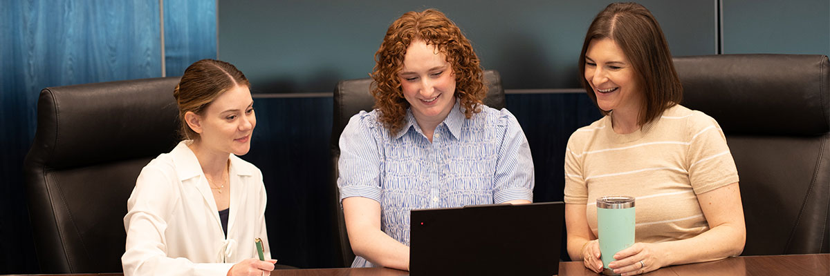 Three people seated around a conference table look at a laptop during a meeting.