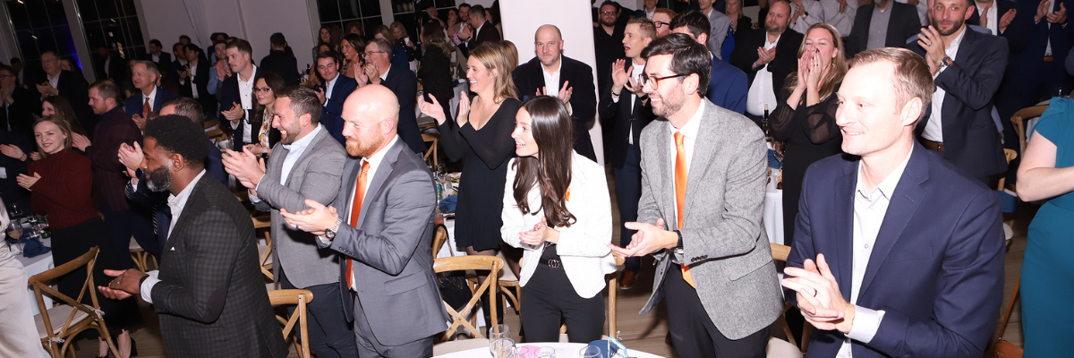 A large group of business professionals stand and applaud at a formal awards event in an indoor venue.