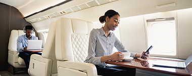 Two people sitting on a private plane, one on laptop and the other on phone. 