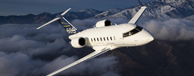 White business jet flying above clouds with mountains in the background.