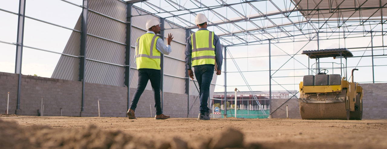 Two construction workers in high-visibility vests and hard hats walk through a large, partially constructed building with a yellow steamroller in the background.