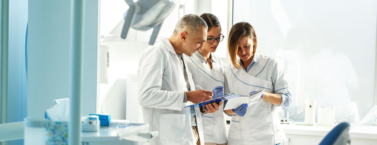 Three clinicians in white coats stand in a bright dental clinic, reviewing documents together while one writes on a clipboard.