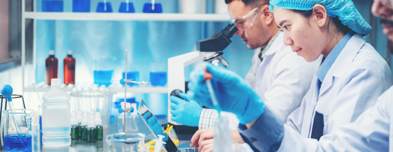 Three scientists in lab coats and protective gear work in a laboratory with various scientific equipment and blue liquid-filled containers.